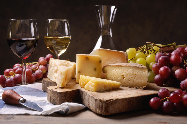 Verschiedene Käse auf einem Holzbrett im Vordergrund, dahinter ein gefülltes Glas Rot- und Weißwein sowie eine Karaffe