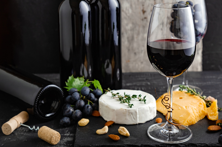 Ein runder Camembert-Käse, daneben ein gefülltes Glas Rotwein und im Hintergrund Rotweinflaschen